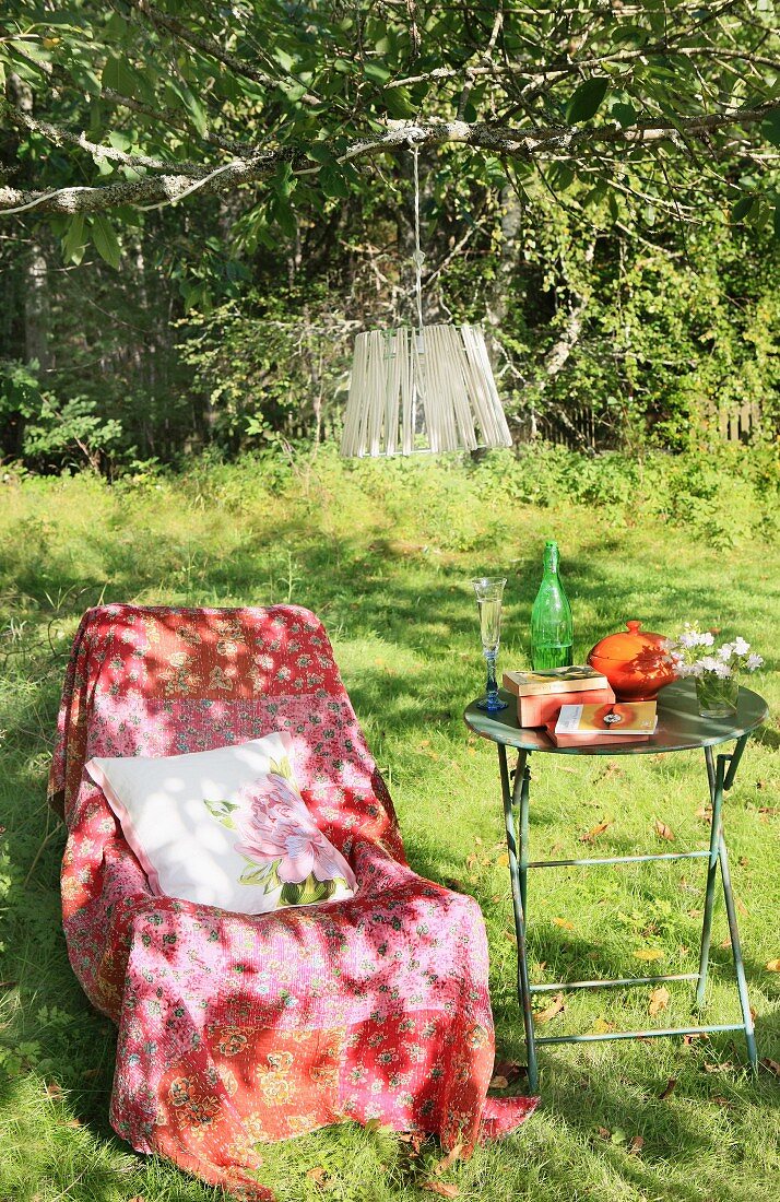 Gemütlicher Stuhl mit Überwurf und ein Gartentisch im Schatten eines Baumes