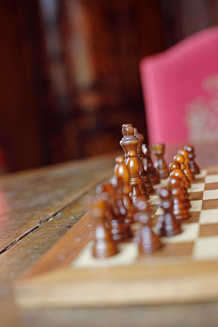 Wooden chess pieces on chessboard on old table with blurred pink chair in background (Schloss Schauenstein)