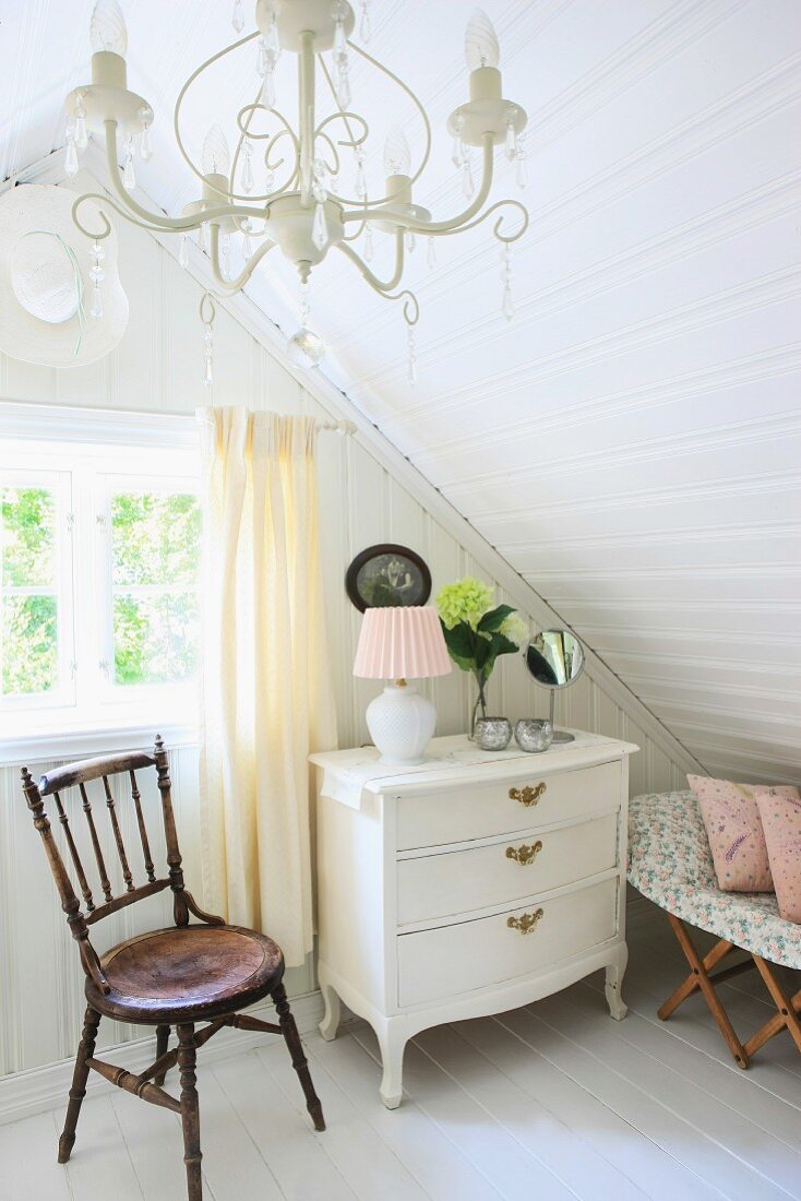 Pale pink table lamp, cosmetic mirror and cut hydrangea on white chest of drawers under sloping ceiling
