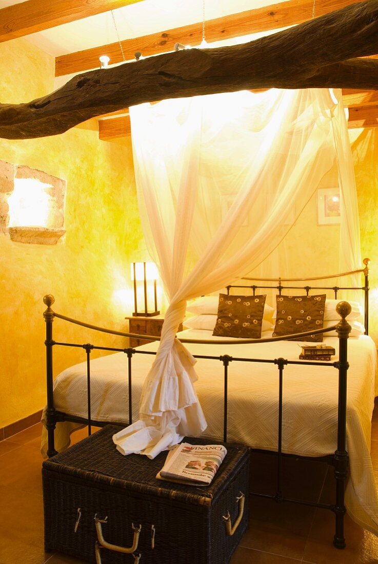 Himmelbett mit Vintage Metallgestell und Truhe aus schwarzem Geflecht in gelbgetöntem Schlafzimmer
