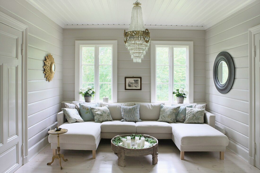 Gemütliche Sitzlandschaft mit Ecksofa in weisses, holzverkleidetes Wohnzimmer mit Kronleuchter und runden Wandspiegeln