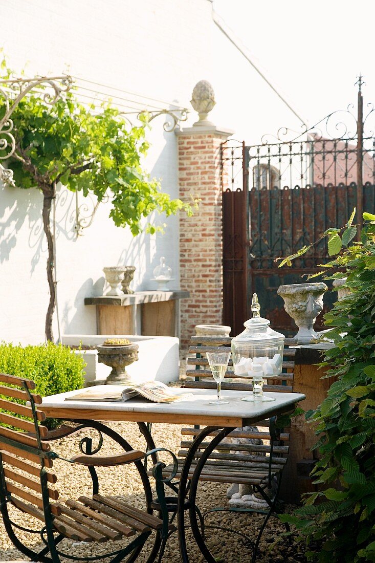 Garden table with marble top and pretty garden chairs next to deciduous hedge; large closed iron gate in background