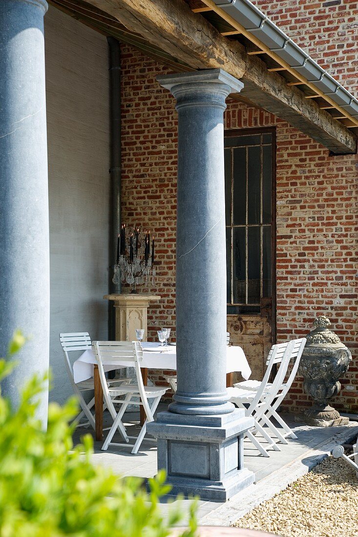 Sonnige Verandaecke mit Kerzendekoration und einfachem Esstisch mit weissen Stühlen; im Vordergrund elegante, graue Säulen