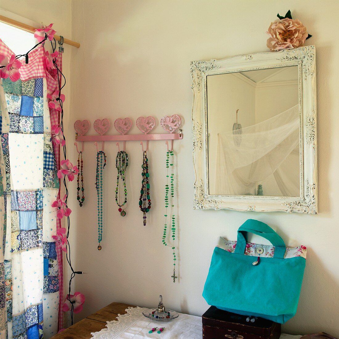 Vintage mirror and strings of beads on pink coat rack with heart motif next to window with patchwork curtain and garland of floral fairy lights