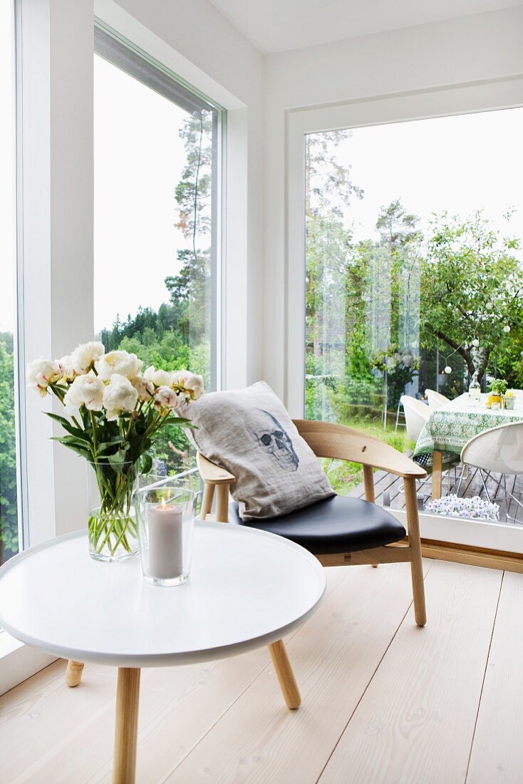 Side table with white, round top and retro wooden chair on pale wooden floor next to large windows