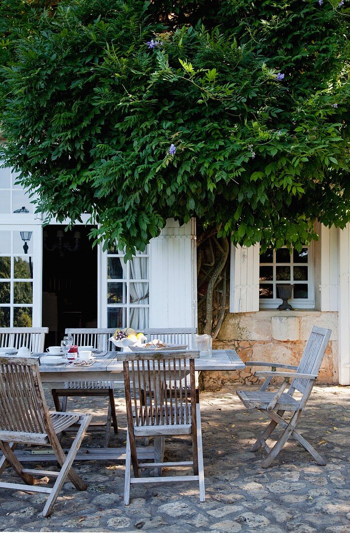 Gedeckter Tisch auf Terrasse mit Natursteinboden vor ländlichem Wohnhaus, teilweise verdeckt von einem Baum