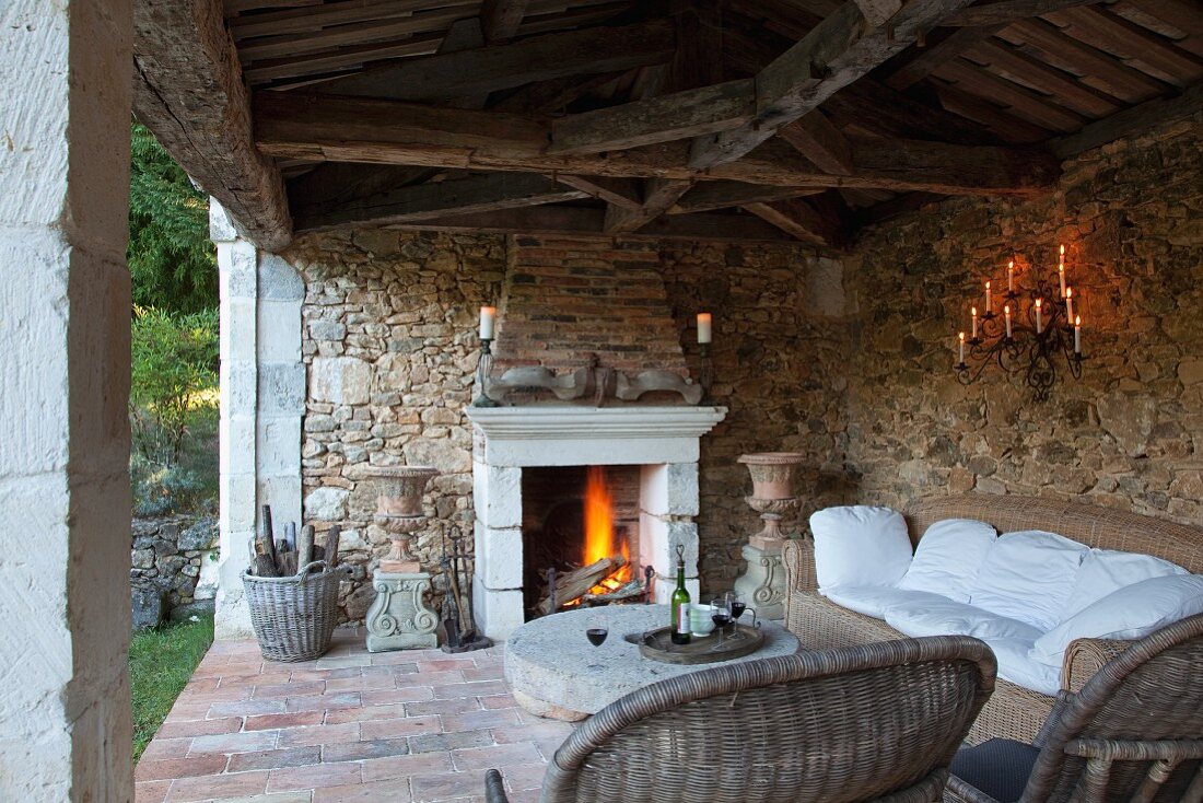 Wicker sofa set on rustic veranda with stone wall and fire in open fireplace