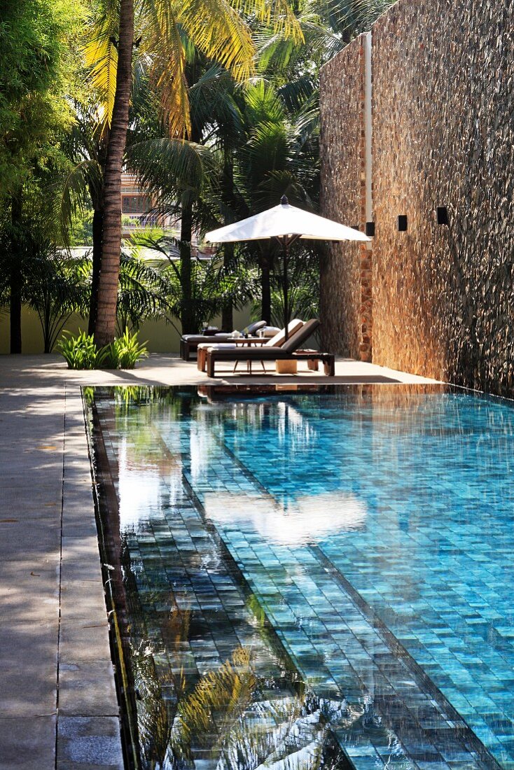Pool and palm trees next to high stone wall; relaxation area with sun loungers and parasol in background