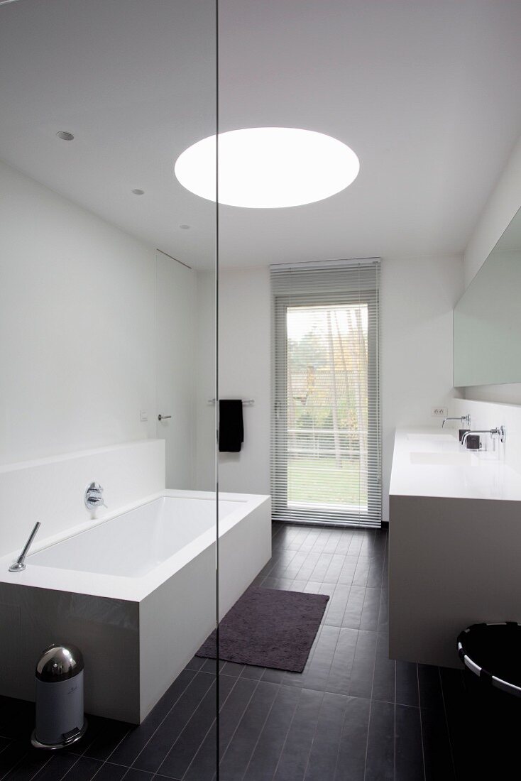 View through glass wall of cubic bathtub, black floor tiles, French door and circular skylight in designer bathroom