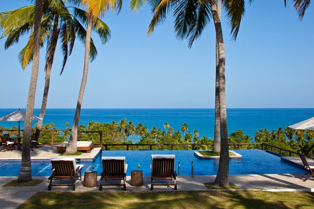 View of blue ocean past pool complex with upholstered wooden loungers below palm trees