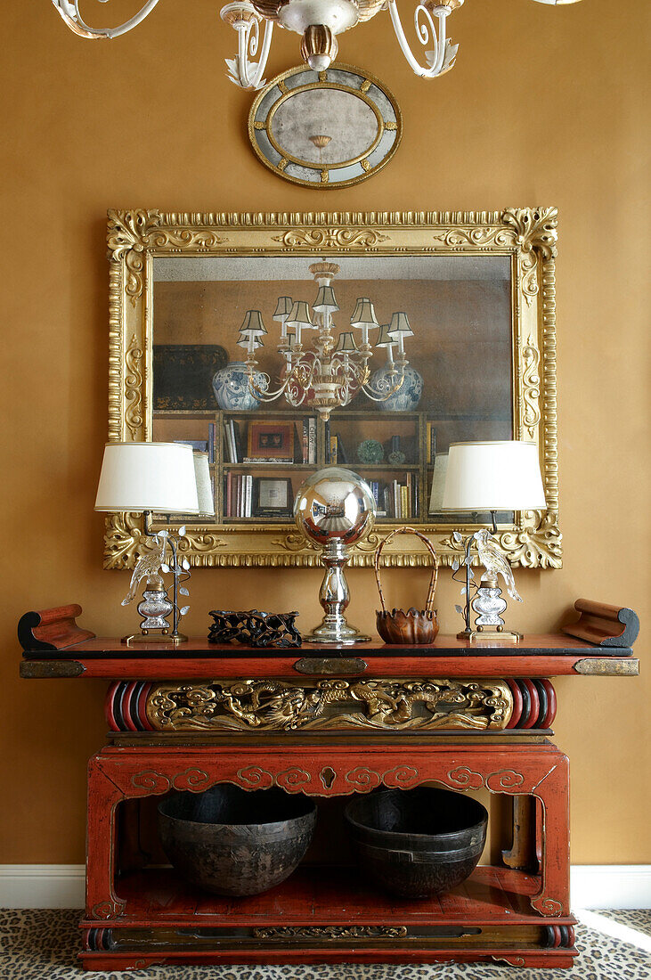 Oriental console table with gilt carvings and two table lamps below gilt-framed mirror on ochre yellow wall