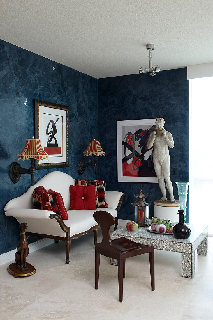 Modern coffee table and antique couch flanked by sconce lamps next to stone sculpture in corner of living room with blue colour-washed walls