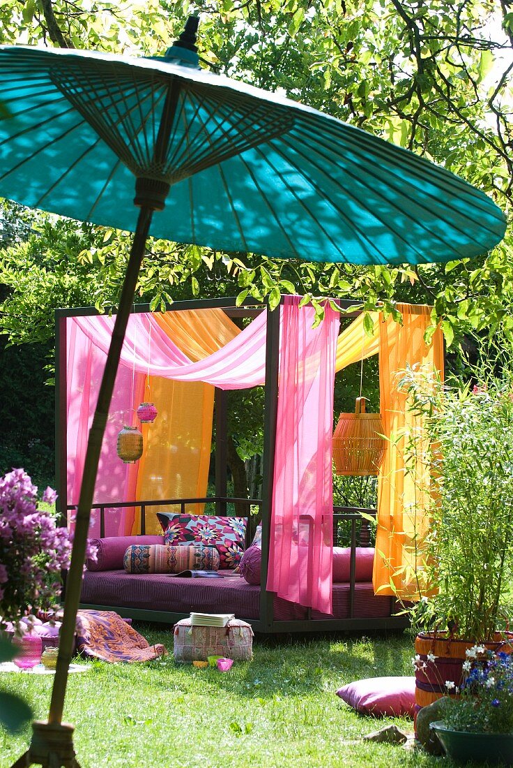 Modern couch with canopy of pink and orange fabrics in sunny garden; Oriental bamboo parasol in foreground