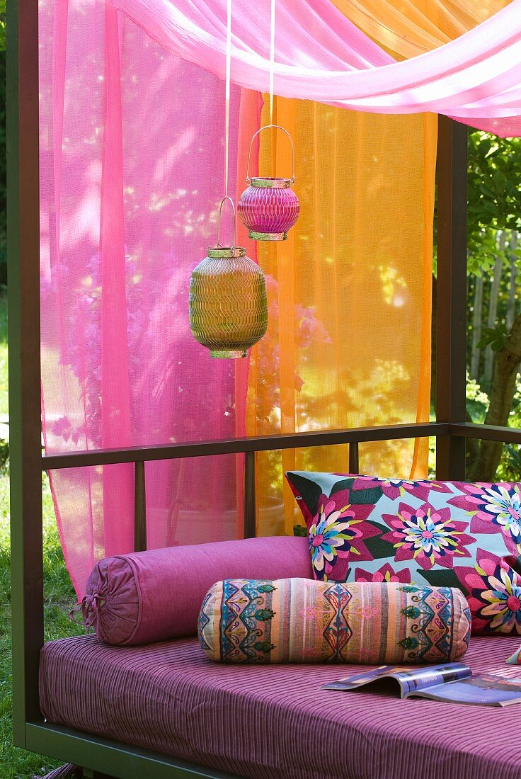 Lampions vor Tüchern in Pink und Orange, bunte Nackenrollen und Kissen als exotische Dekoration auf modernem Tagesbett