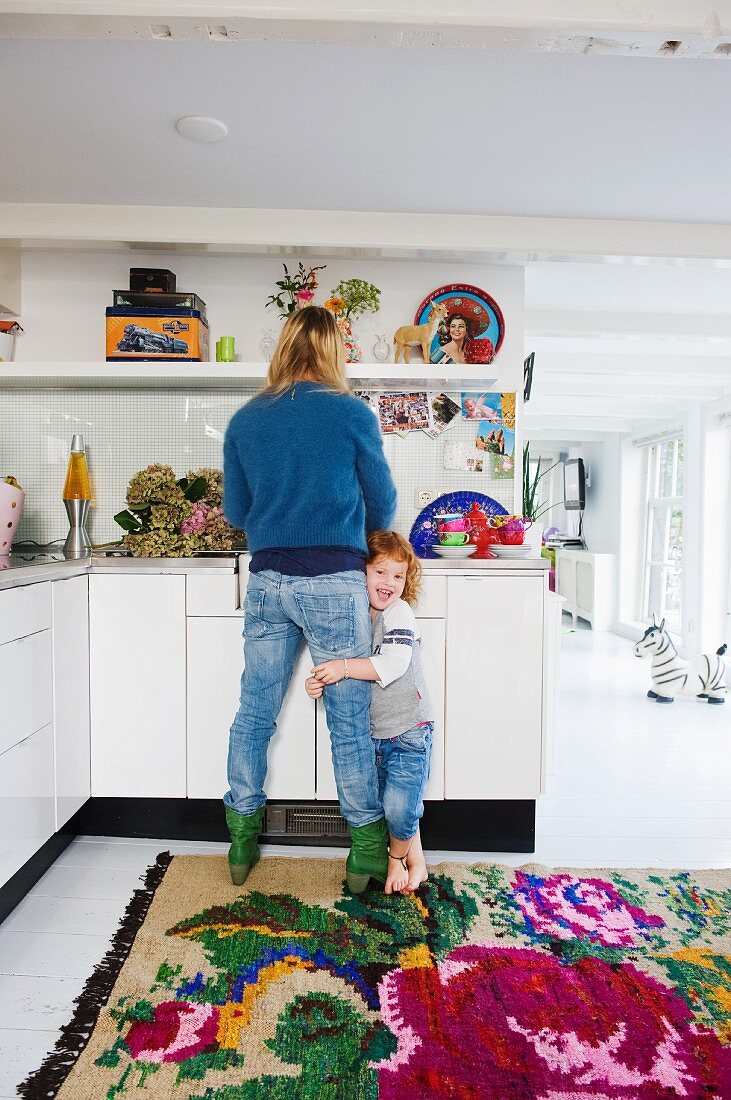 Mother working in kitchen with child hanging on leg; white corridor with colourful rug on floor