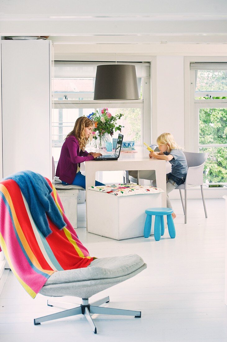 Two children with laptop and toys sitting in cool, white dining area; colourful striped towel on swivel chair in foreground