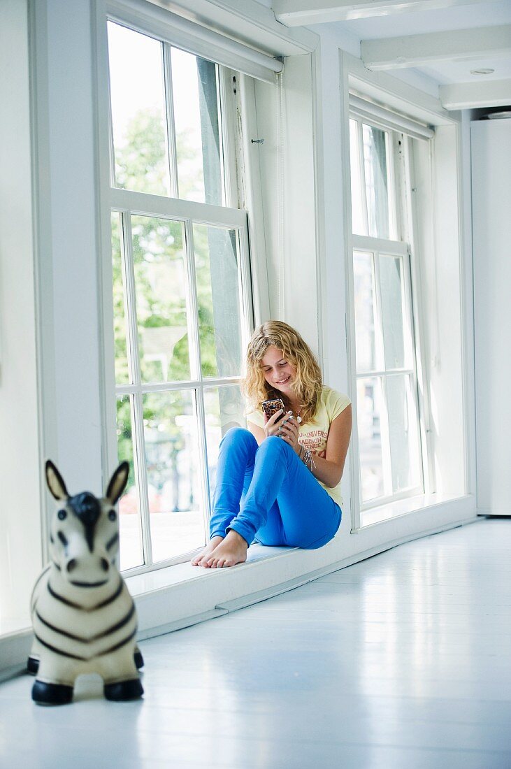 Teenage girl sitting on window sill in white interior; toy animal in foreground