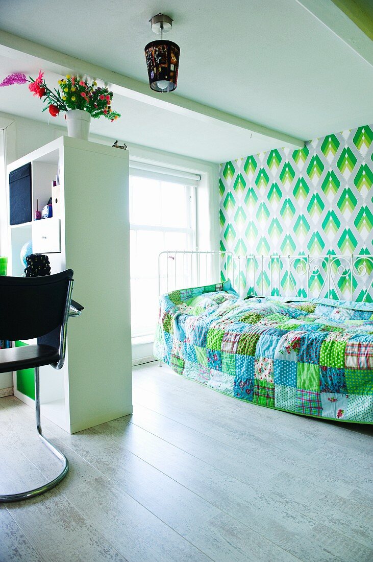 Child's bedroom with shelving as partition between bed and desk; shades of blue and green in patchwork blanket and retro wallpaper