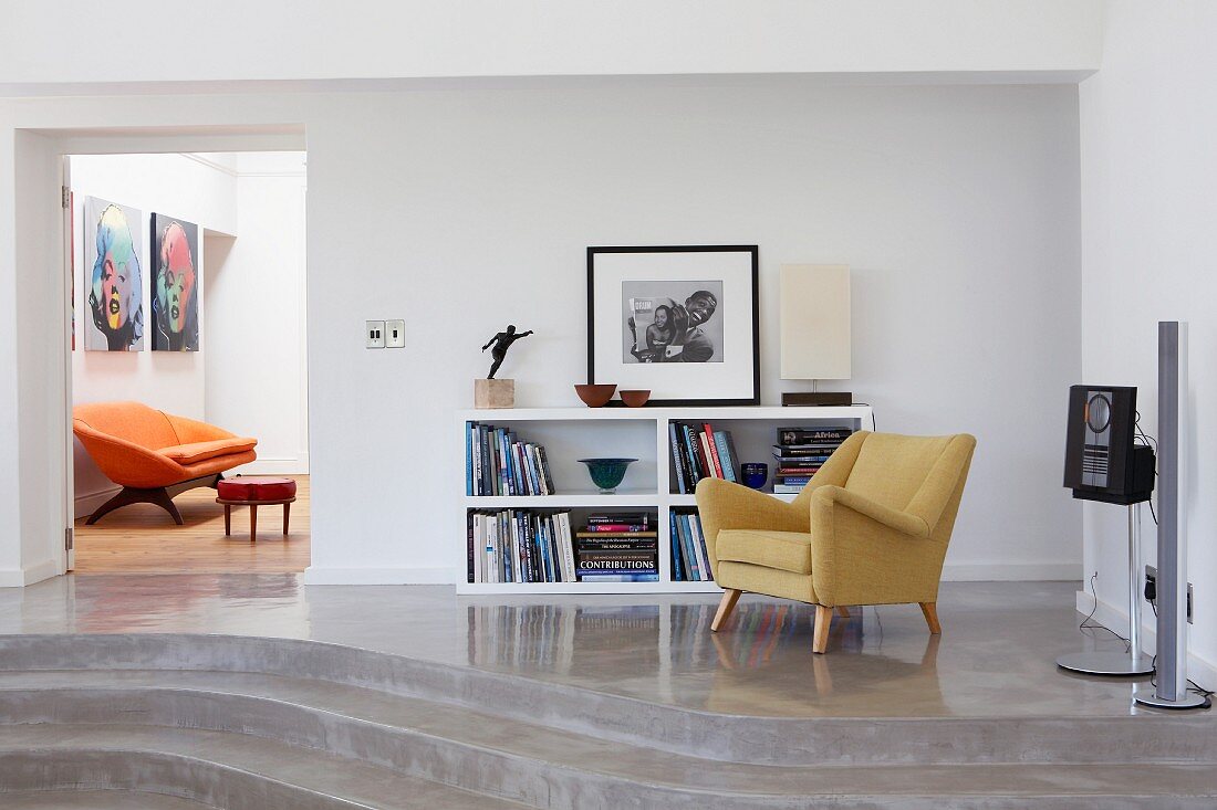 50er Jahre Sessel und halbhohes Regal auf Podest mit Stufen aus Beton und Blick durch offene Tür auf orangefarbenes Sofa