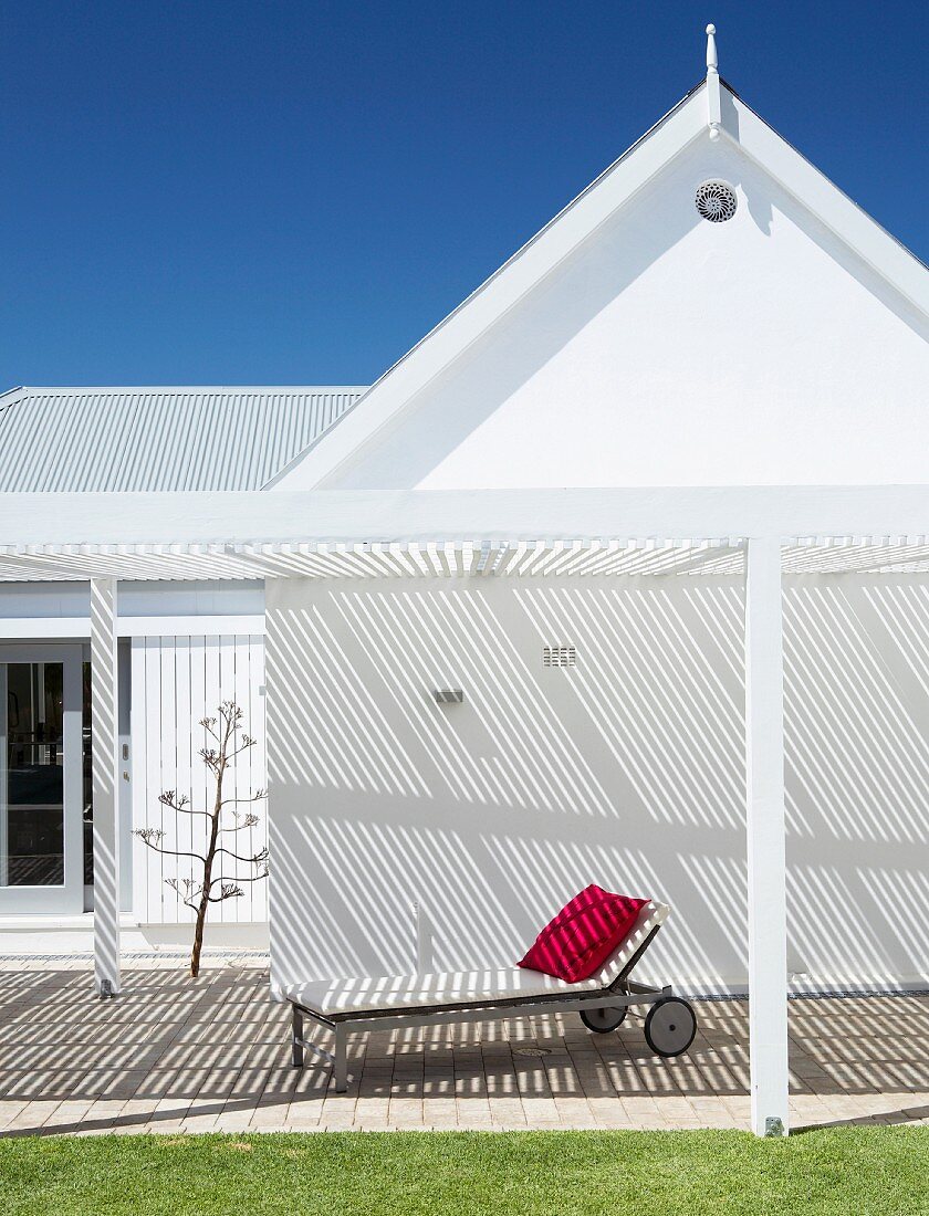 Sun lounger under white pergola attached to gable facade of angular house