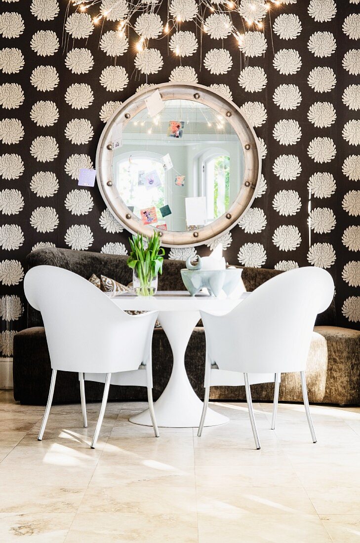 weiße Kunststoffstühle (Phillipe Starck) mit weißem Bistrotisch vor braunem Sofa und kreisfömiger Spiegel an Wand mit gemusterter Tapete