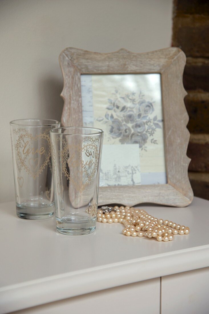 weiße Perlenkette, zwei Gläser und Bild im Holzrahmen auf einer Kommode
