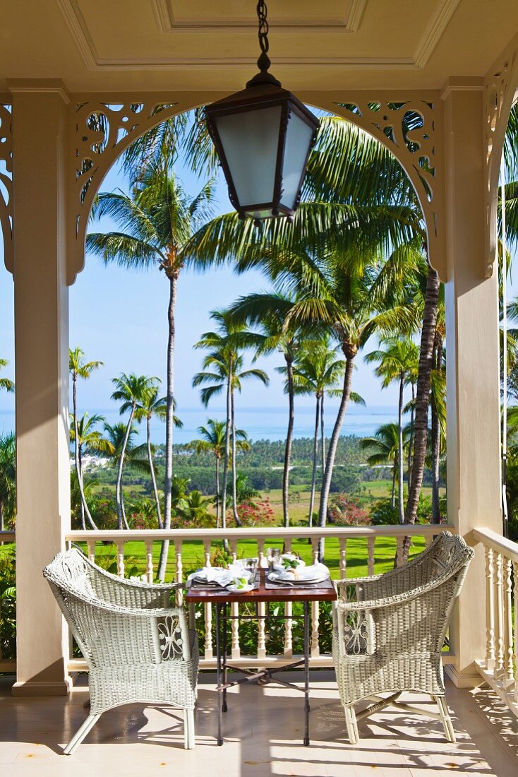weiße Rattanstühle um gedeckten Tisch auf Veranda mit im Kolonialstil und Blick in Palmengarten