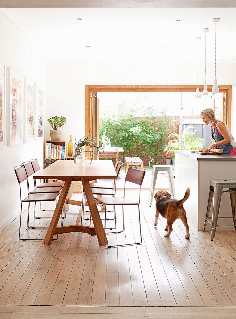 Filigrane Stühle mit Chromgestell an Holztisch gegenüber Theke und Frau bei Küchenarbeit in modernem Ambiente mit offener Terrassentür