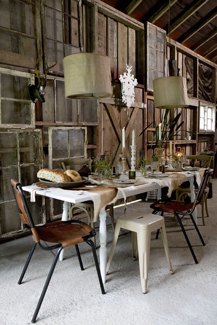 Festlich gedeckter Tisch unter Hängeleuchten vor improvisierter Holzwand aus Fensterrahmen und Paneelen in scheunenartigen Raum