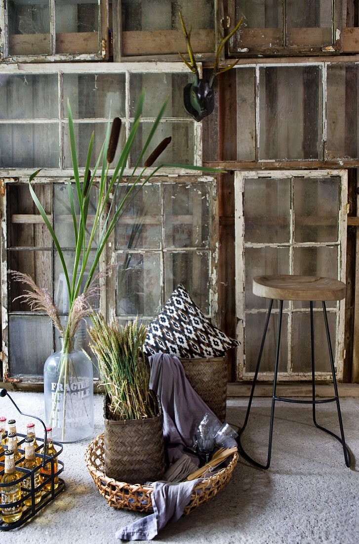 Stillleben aus Körben mit Pflanzendeko neben Barhocker vor improvisierter Wand mit Vintage Fenstern vor Holzwand