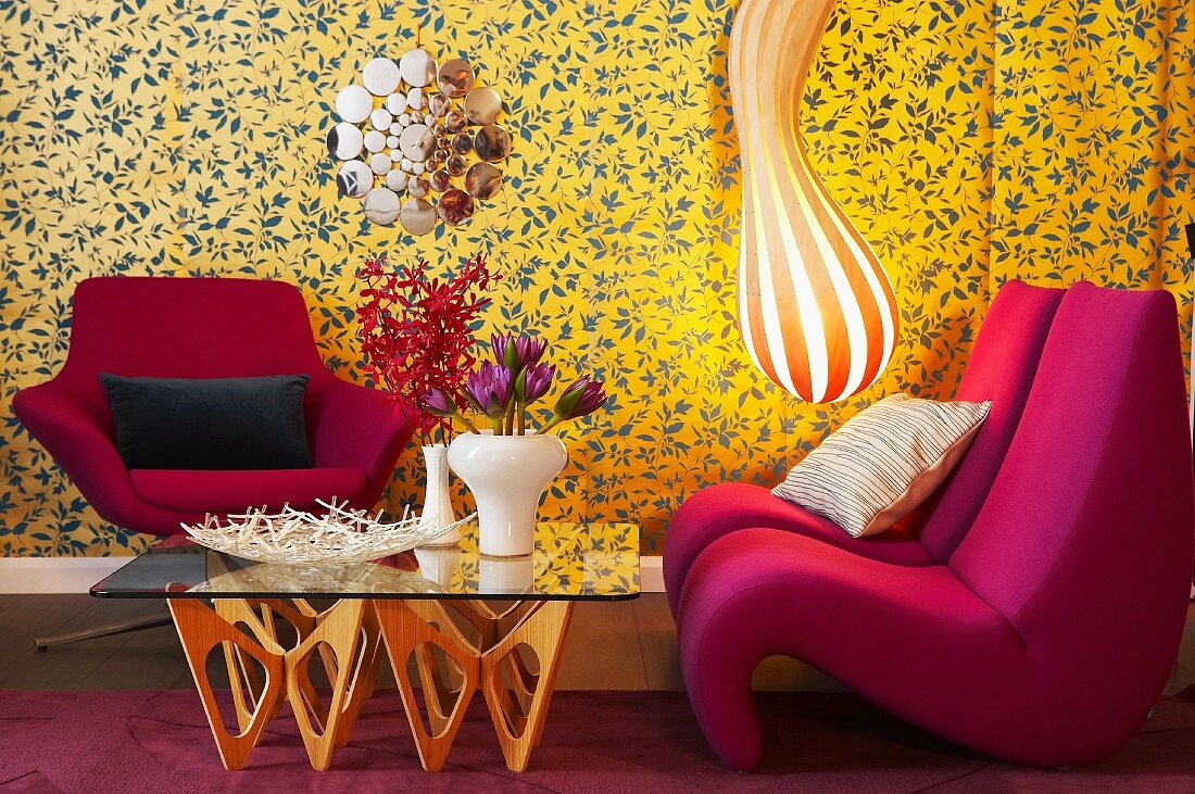 Gestreifte Designer-Lampe, Sessel und Glastisch vor gemusterter Tapete in Wohnzimmerecke