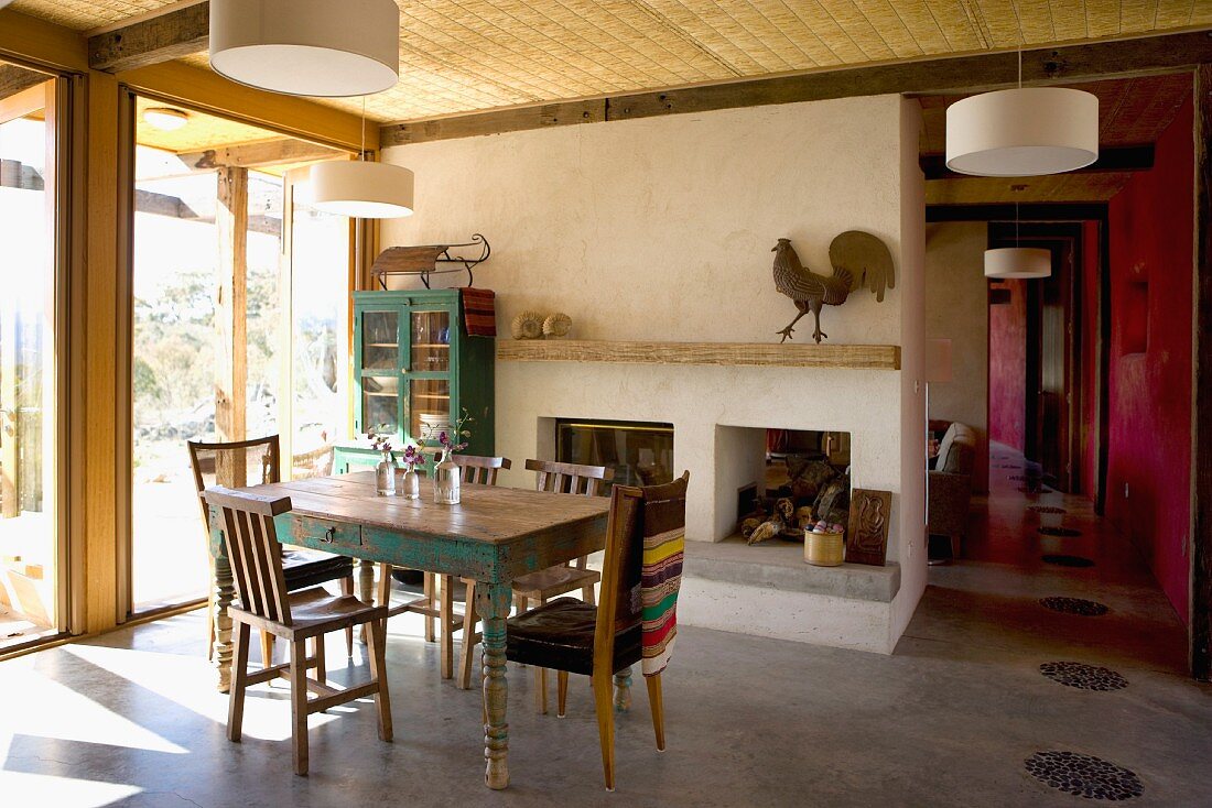 Open, minimalist dining area with assorted chairs around a rustic kitchen table in front of a window wall and an open fireplace