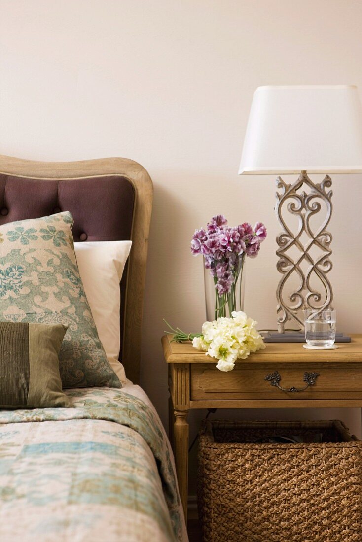 Wooden bedside table and table lamp with white lampshade and metal base next to bed with upholstered headboard