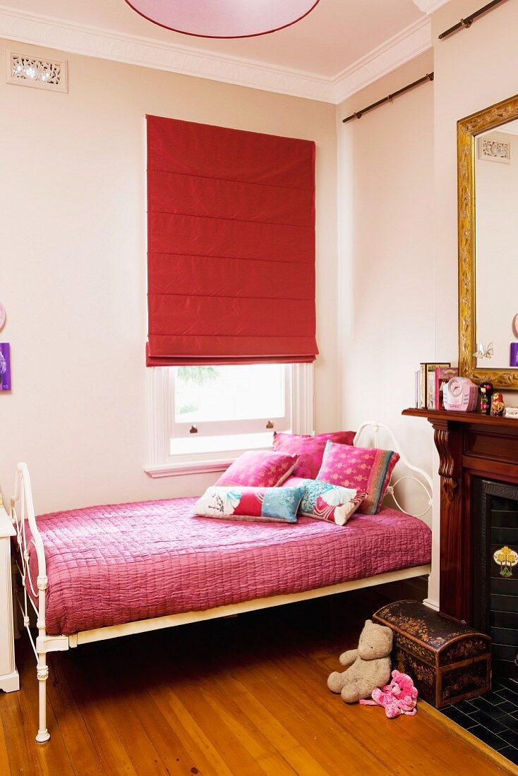 Girl's bedroom with antique, white metal bed frame below window with closed, red Roman blind and open fireplace in traditional interior