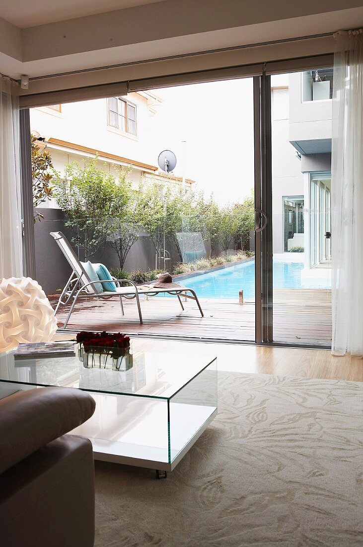 Cubic, glass coffee table on pale rug in front of open sliding door and view across terrace to pool in courtyard