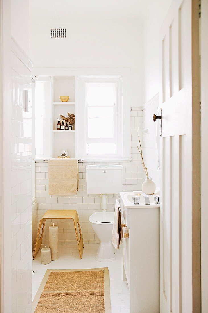View through open door of modern wooden stool next to toilet below window in white-tiled bathroom