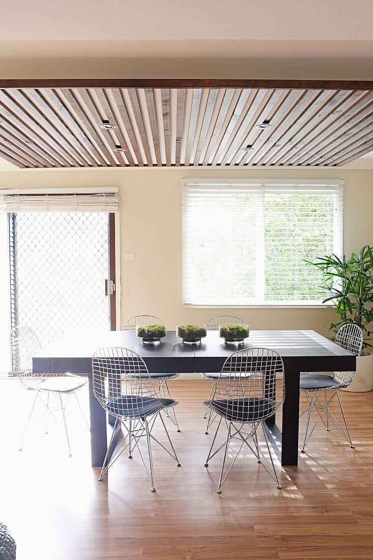 Sunny dining area with black table and wire chairs with seat cushions below suspended ceiling