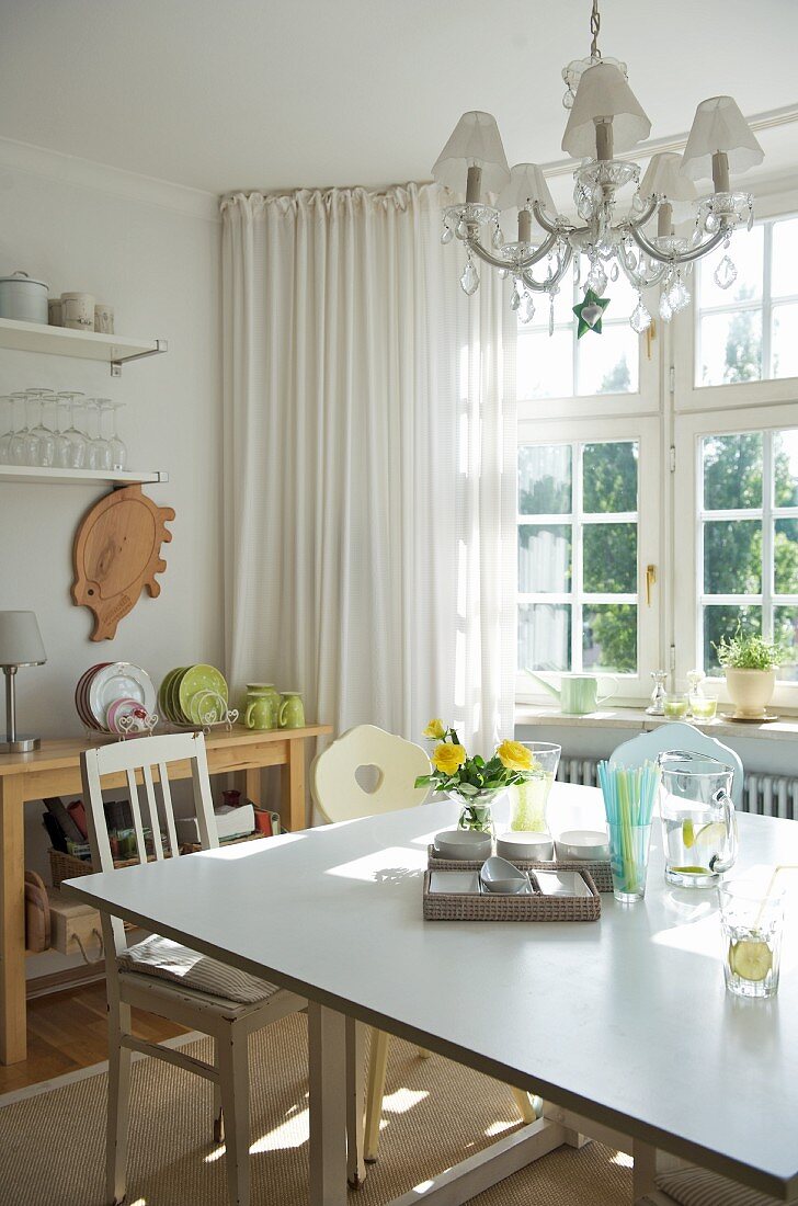 Sunny, simple kitchen with bay window, square dining table and vintage chairs; crystal chandelier above table