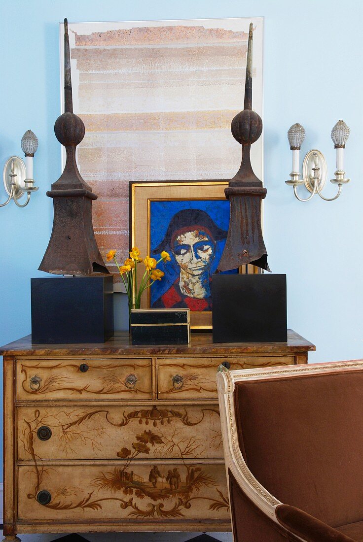 Teilweise sichtbarer Stuhl vor Kommode mit Intarsien und Kunstobjekte vor Bildern an blau getönter Wand