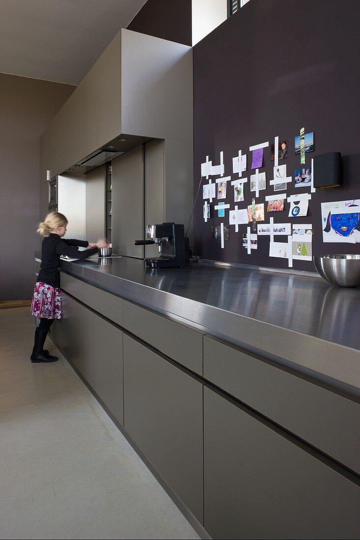 Mädchen an der langen Arbeitsfront einer Designerküche aus grauem HPL und Edelstahl; angepinnte Erinnerungsstücke an der Wand
