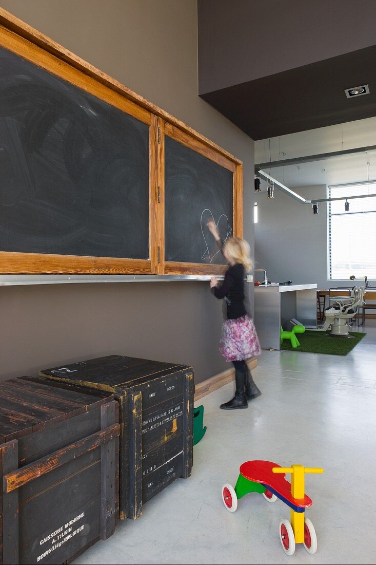 Offener Wohnraum mit alter Wandtafel in umgenutztem Schulhaus; alte Transportkisten und buntes Holzrad auf poliertem Betonboden
