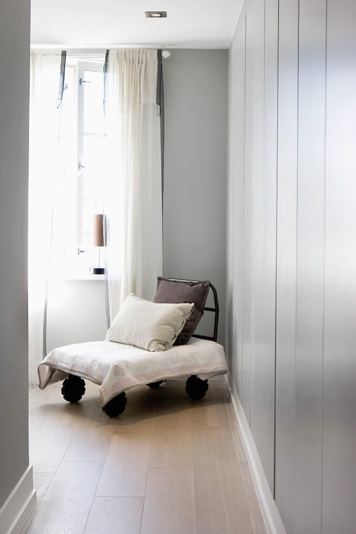 Selbstgebauter Sessel mit Kissen auf Holzboden vor grauem Einbauschrank und Fenster mit luftigem Vorhang