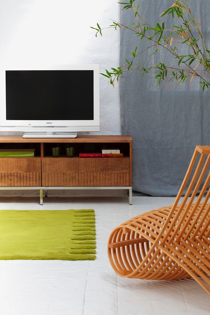 Moderner Stuhl aus gebogenen Holzleisten vor grünem Teppichläufer auf weißem Fliesenboden und Lowboard mit Flachbildschirm