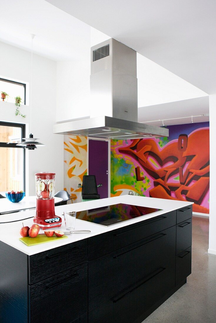 Modern, free-standing kitchen counter with black wooden fronts and stainless steel extractor hood in front of graffito mural on wall
