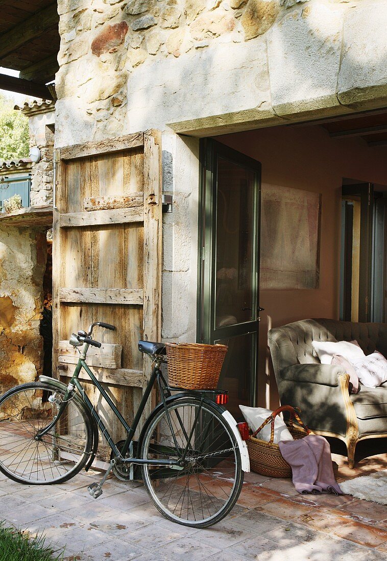 Fahrrad mit Korb vor rustikalem Natursteinhaus mit Türlade und Blick durch offene Terrassentüren auf Polstersofa