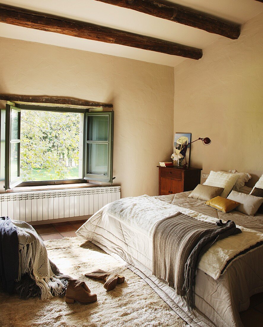 Comfortable bed with blankets and scatter cushions in front of open window in bedroom with wood-beamed ceiling