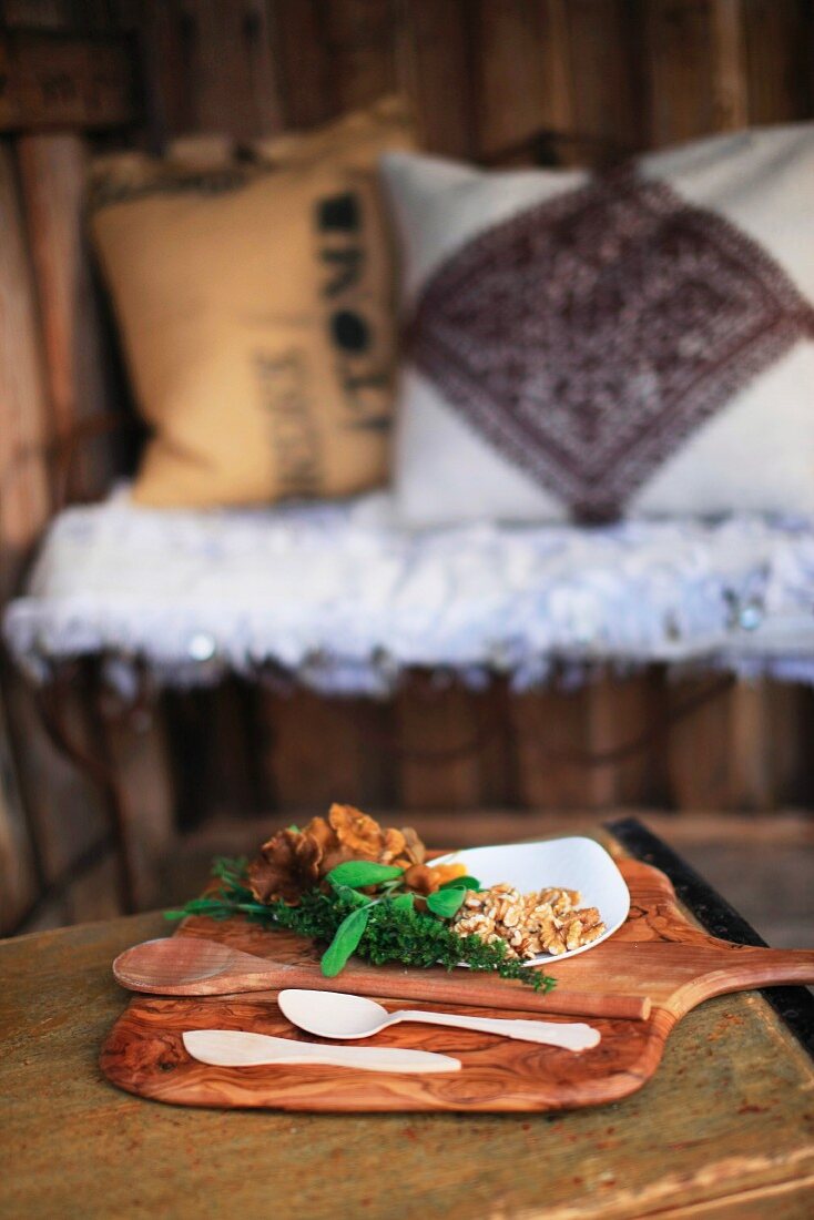 Chanterelles, walnuts, herbs and wooden spoons on wooden board; cushions on wire bench in blurred background