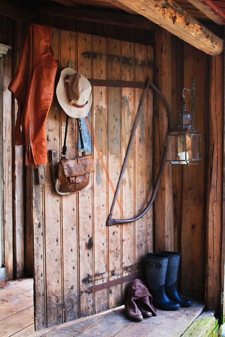 An Brettertür eines Holzunterstandes aufgehängte Lederjacke und Damenhut neben Baumsäge und Laterne