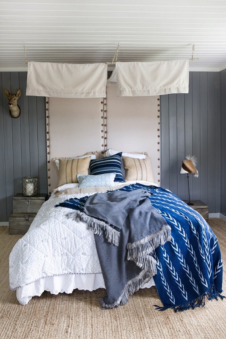 Different coloured blankets on double bed with canopy and upholstered headboards on grey wooden wall in rustic bedroom