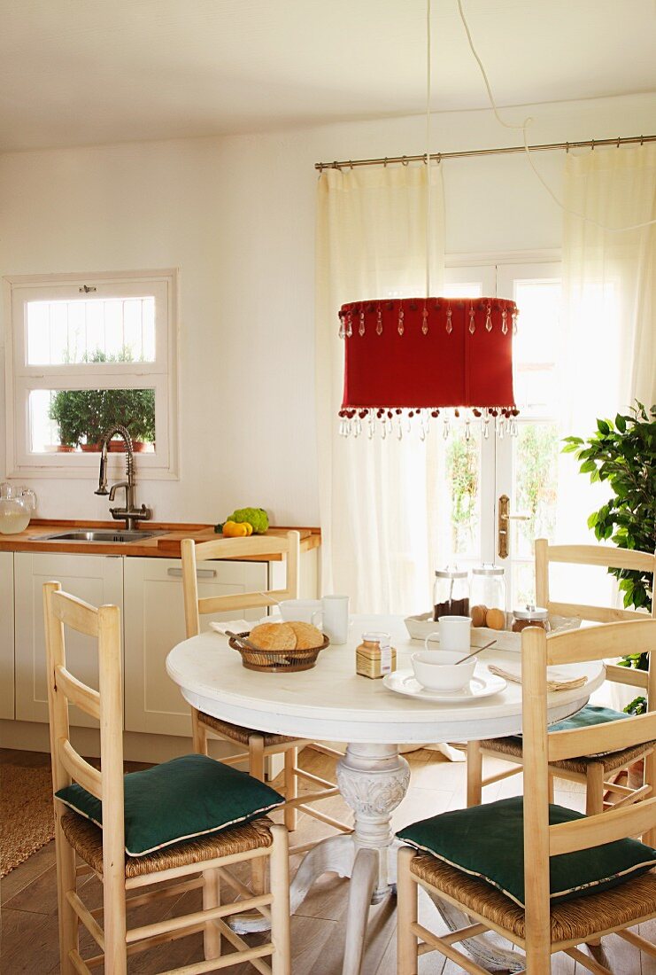 Mediterrane Holzstühle um gedeckten Frühstückstisch unter Hängeleuchte mit rotem Schirm vor Terrassentür in weißer Küche